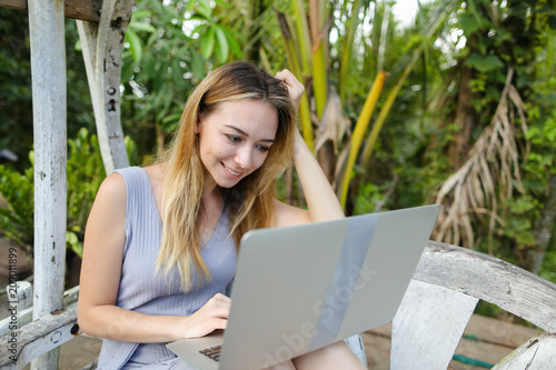 Happy blonde woman using laptop and sitting in exotic garedn with palms in background. Concept of summer vacations and modern technology. photo