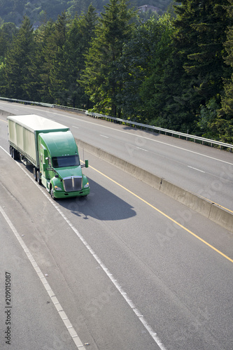 Green big rig American semi truck with tented semi trailer moving on wide divided highway with trees on background