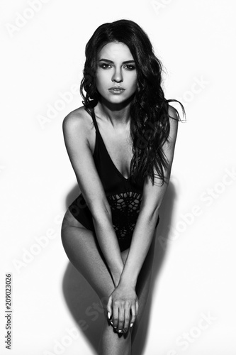Young sexy slim tanned woman in black swimsuit posing against white background. Black and white fashion portrait of beautiful girl with long wavy brunette hair. Swimwear or bikini model.