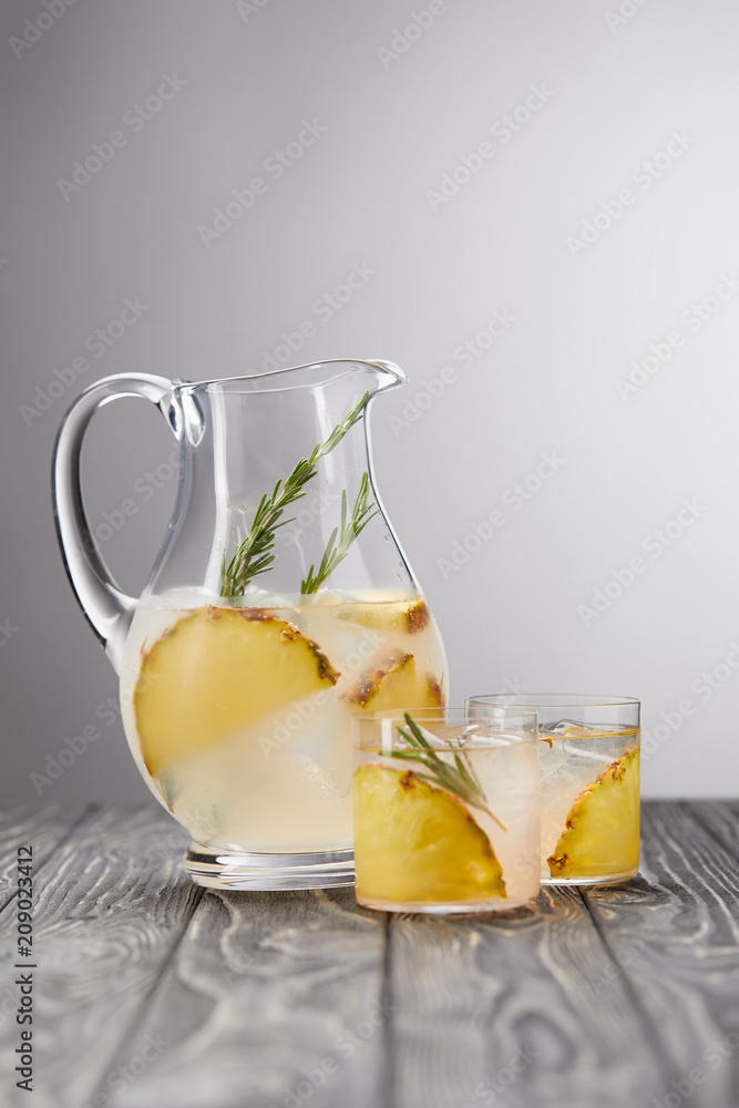 jug and two glasses of lemonade with pineapple pieces, ice cubes and rosemary on grey wooden tabletop jug and two glasses of lemonade with pineapple pieces, ice cubes and rosemary on grey wooden tabletop
