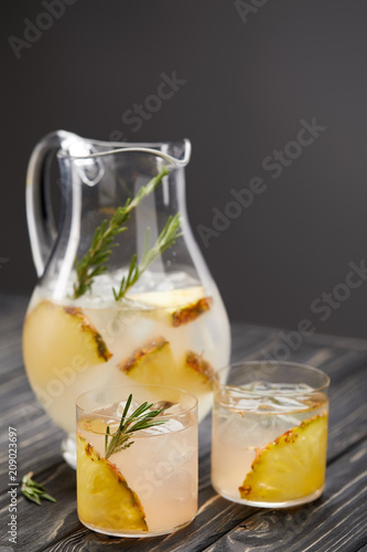 jug and two glasses of lemonade with pineapple pieces, ice cubes and rosemary on grey wooden tabletop