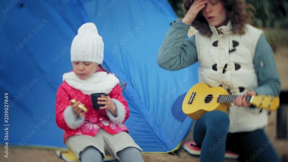 A beautiful girl with her family plays on a ukulele on the river bank near a tourist tent A beautiful girl with her family plays on a ukulele on the river bank near a tourist tent