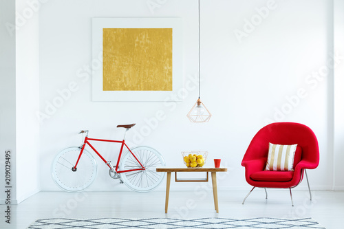 Real photo of a bright living room interior with a red armchair, wooden table with lemons in the basket, lamp, bike and painting on the wall