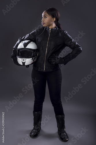 Black female motorcycle biker or race car driver or stuntwoman wearing leather racing suit and holding a protective helmet. She is standing confidently in a studio