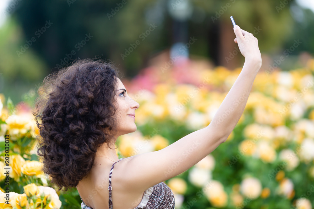 A portrait of Caucasian woman with dark curly hair taking selfie near yellow rose bushes in a rose garden A portrait of Caucasian woman with dark curly hair taking selfie near yellow rose bushes in a rose garden