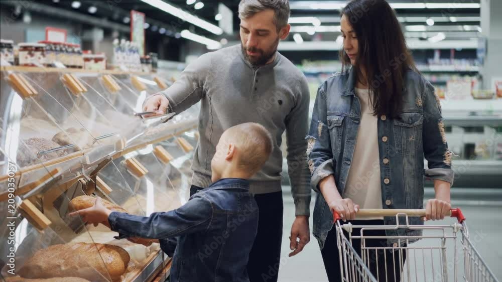 Helpful son is helping his parents to buy bread , father is opening container and boy is taking loaf and smelling it then putting in trolley. Shopping together concept. Helpful son is helping his parents to buy bread , father is opening container and boy is taking loaf and smelling it then putting in trolley. Shopping together concept.