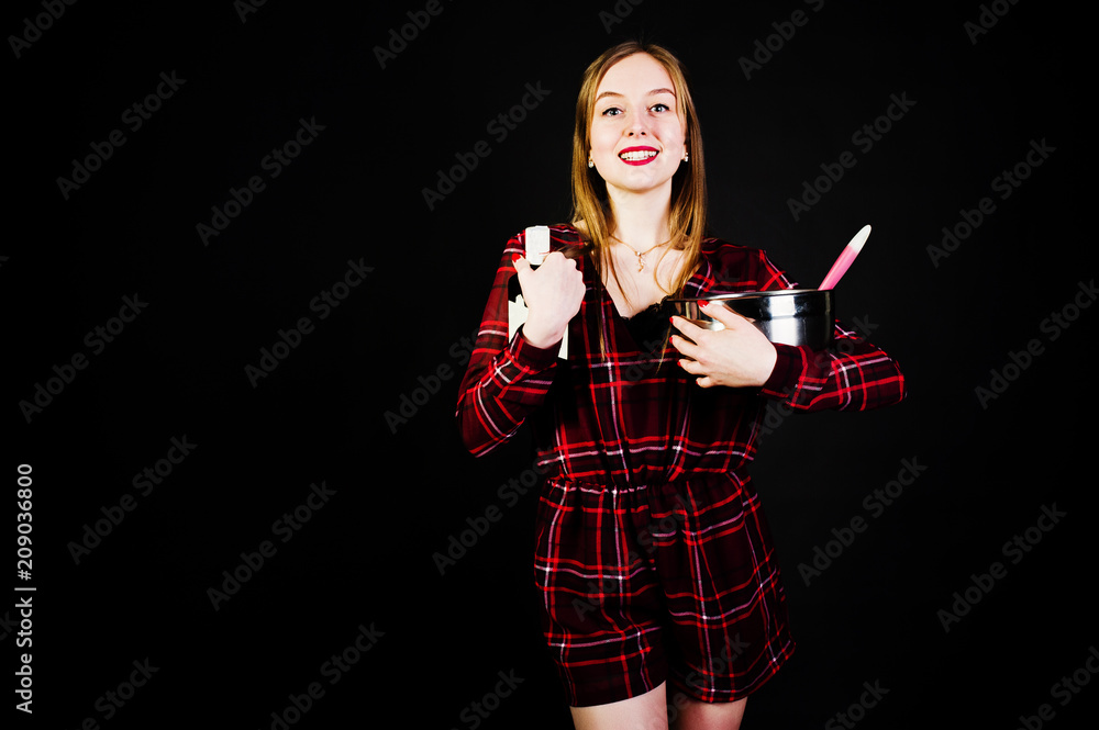 Young housewife in checkered dress with saucepan and kitchen spoon and bottle isolated on black background. Young housewife in checkered dress with saucepan and kitchen spoon and bottle isolated on black background.