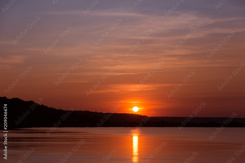 red sunset, the sun is reflected in the water, light fluffy clouds, black coastline, concept nature and summer