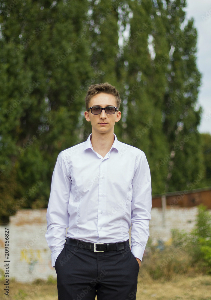 Young guy in white shirt and black trousers. Handsome smart man in glasses standing outside with green trees on background. Business portrait. Vertical photo. Young guy in white shirt and black trousers. Handsome smart man in glasses standing outside with green trees on background. Business portrait. Vertical photo.