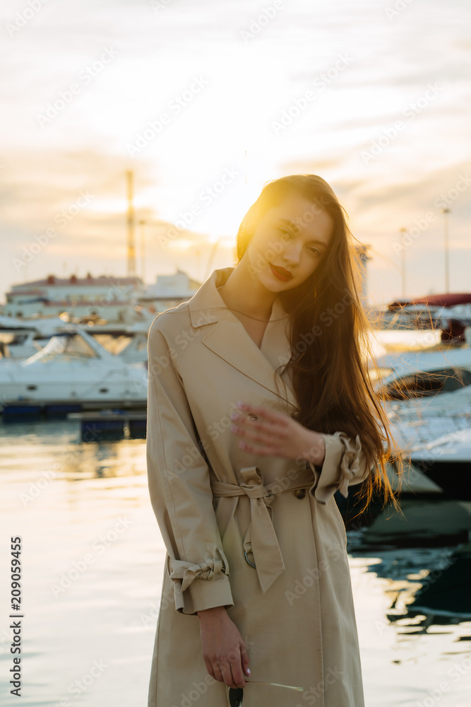 stylish long-haired girl in fashionable beige coat posing in seaport at sunset
