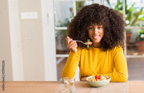 African american woman eating pasta salad at home with a happy face standing and smiling with a confident smile showing teeth