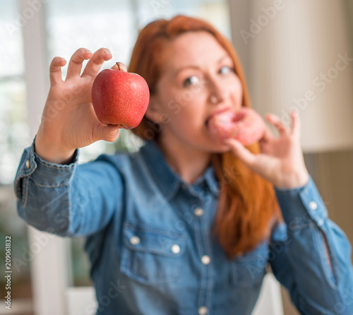 Redhead woman chooses between apple and donut with a happy face standing and smiling with a confident smile showing teeth photo