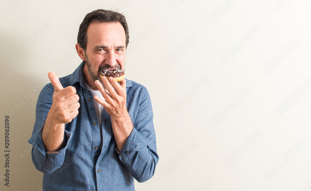 Senior man eating chocolate donut happy with big smile doing ok sign, thumb up with fingers, excellent sign Senior man eating chocolate donut happy with big smile doing ok sign, thumb up with fingers, excellent sign