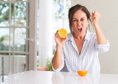 Middle aged woman holding orange fruit annoyed and frustrated shouting with anger, crazy and yelling with raised hand, anger concept