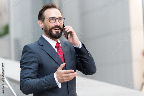 A good deal! Bearded businessman speaks by phone and laughs.View of a Young attractive business man in glasses using smartphone. Smiling formal dressed businessman talking on cell phone with customers