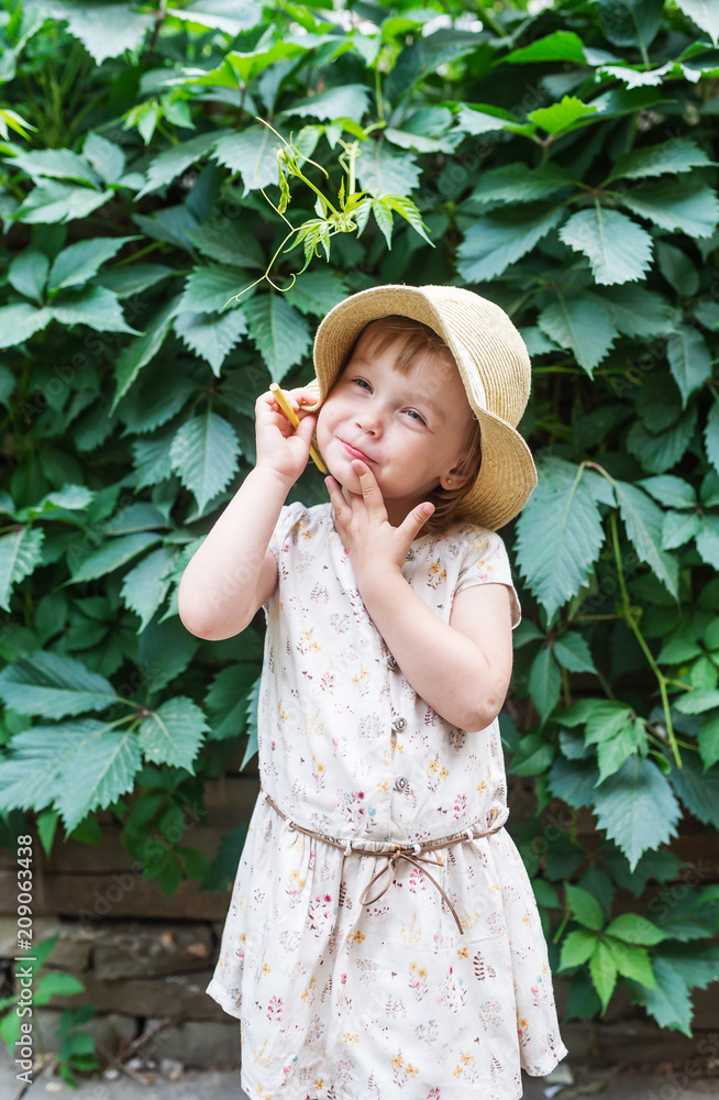 Little girl in straw hat with yellow pencil on background green plant leaves in garden summer.