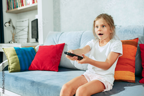 Little casual girl watching tv at home. Female kid sitting on sofa with TV remote and switching channels