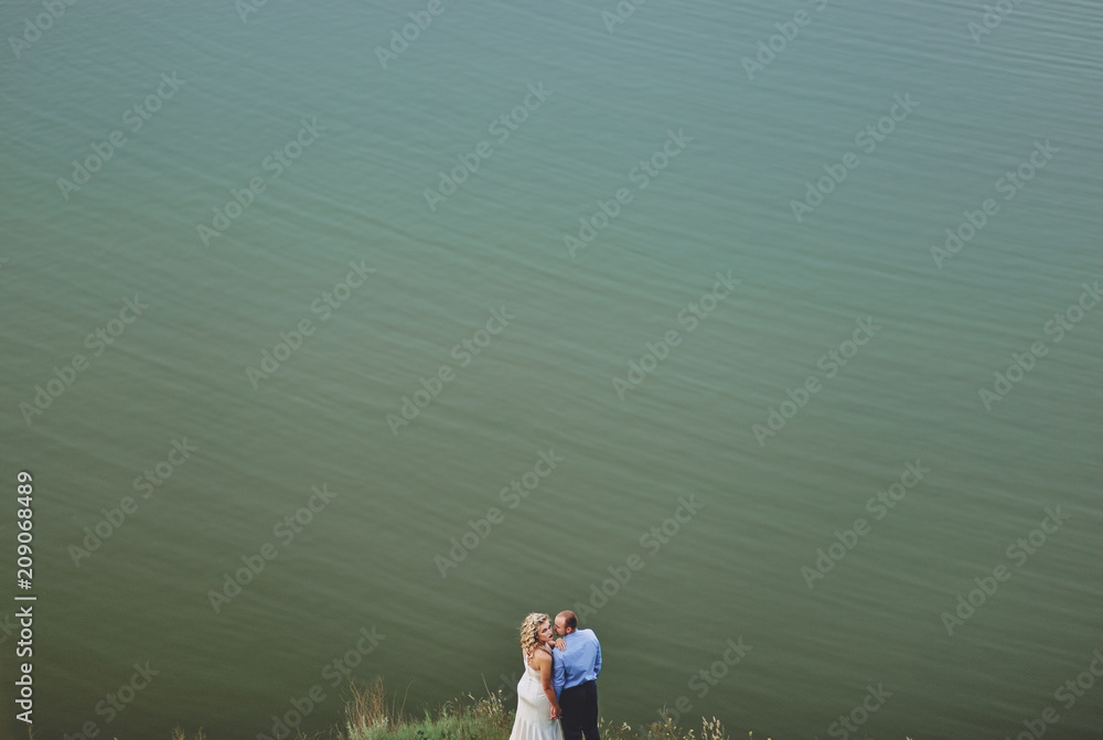 Fototapeta premium Stylish newlyweds stand on the background of the lake. A young groom and a beautiful bride with curly hair stand against the sea and embrace. Wedding.