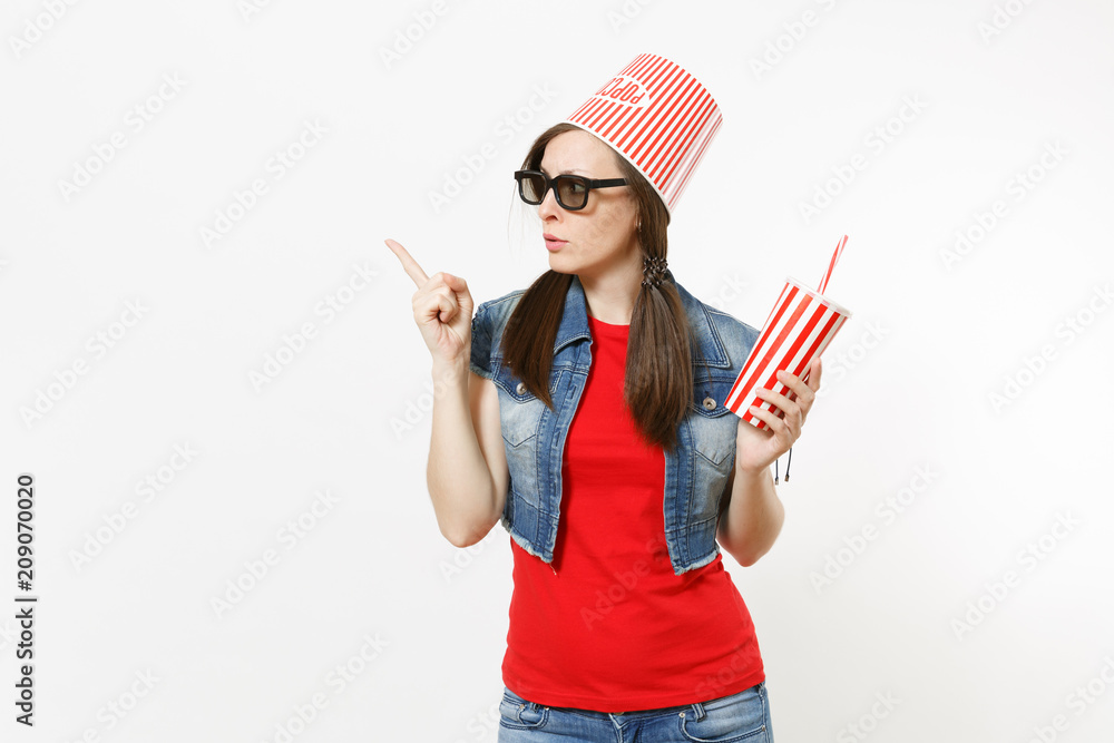Young concerned woman in 3d glasses with bucket for popcorn on head watching movie film holding cup of soda or cola pointing index finger on copy space isolated on white background. Emotions in cinema Young concerned woman in 3d glasses with bucket for popcorn on head watching movie film holding cup of soda or cola pointing index finger on copy space isolated on white background. Emotions in cinema