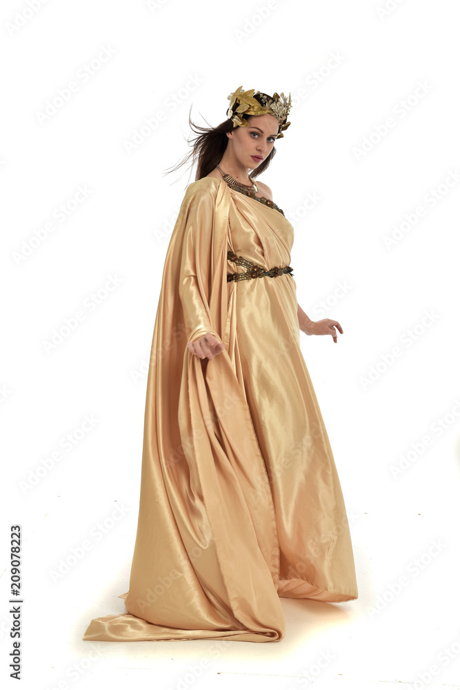 full length portrait of brunette woman wearing long golden grecian gown. standing pose on white studio background.