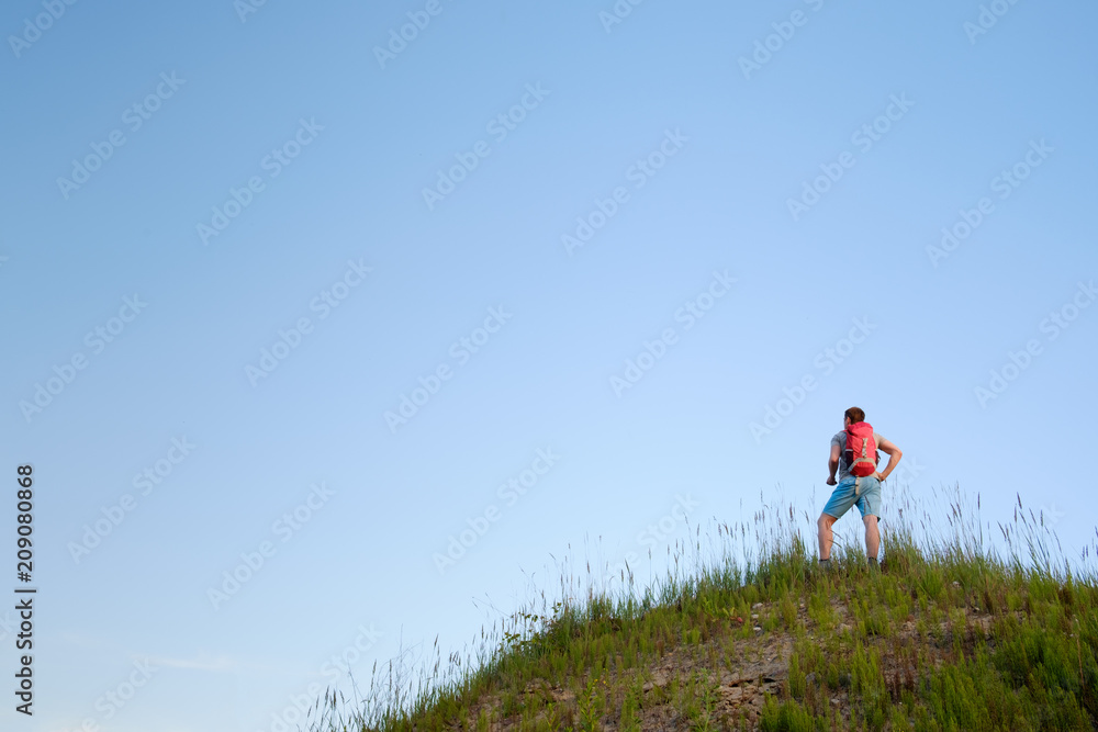 traveler with orange backpack hiking in the hills on blue sky background, view from behind