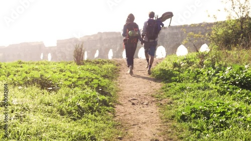 Young lovely couple backpackers tourists walking holding hands toward roman aqueduct arches in parco degli acquedotti park ruins in rome on romantic misty sunrise with guitar and sleeping bag slow photo