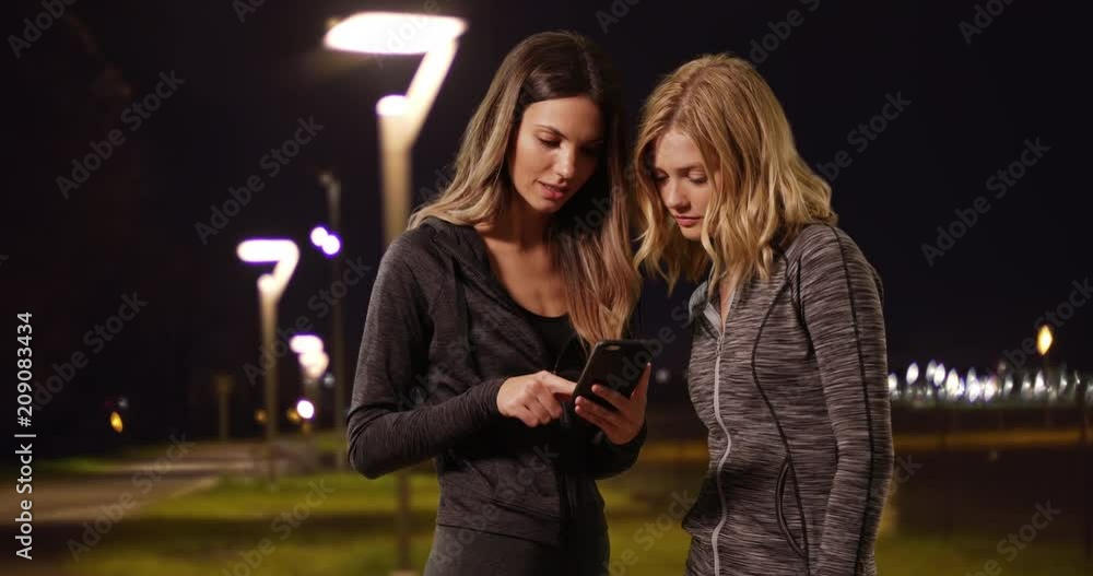 Healthy cute white girls looking at stopwatch and smart phone while out on a night jog, Two attractive young women timing jog with stop watch and smartphone outside at night, 4k Healthy cute white girls looking at stopwatch and smart phone while out on a night jog, Two attractive young women timing jog with stop watch and smartphone outside at night, 4k