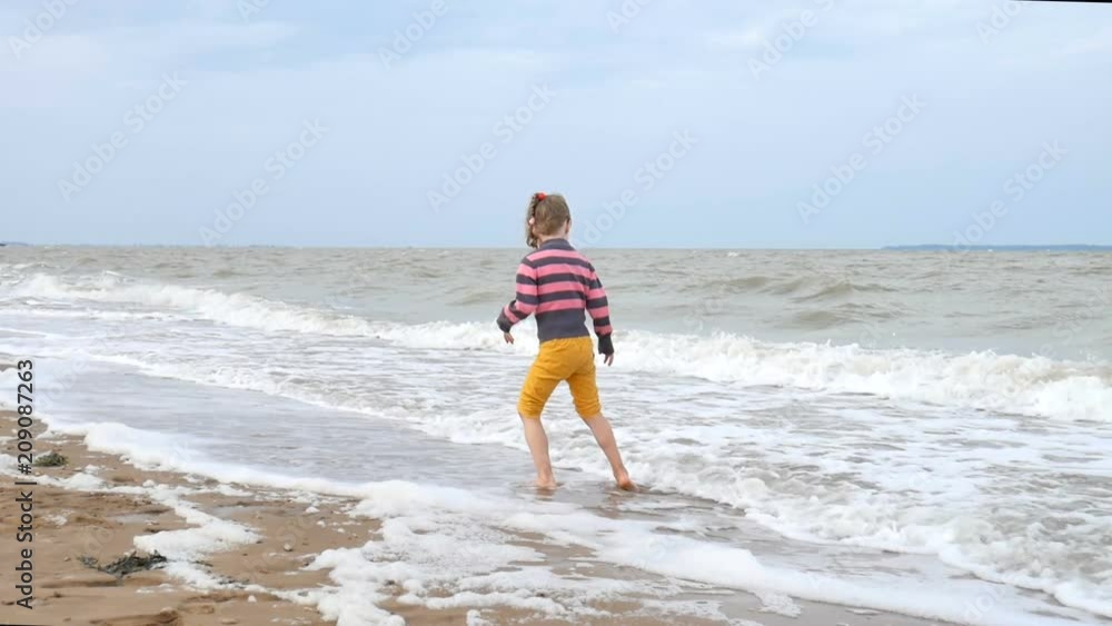 Happy Active Girl Playing with Waves on the Beach Near the Water. A Weak Storm at The Sea, Strong Wind. Summer Vacation, Happy Childhood Concept. Happy Active Girl Playing with Waves on the Beach Near the Water. A Weak Storm at The Sea, Strong Wind. Summer Vacation, Happy Childhood Concept.