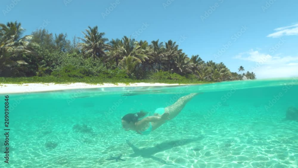 SLOW MOTION, HALF UNDERWATER: Bright summer sun shines on Caucasian woman diving past a black starfish on the sandy ocean floor. Active female on holiday swims along sandy beach in the glassy ocean. SLOW MOTION, HALF UNDERWATER: Bright summer sun shines on Caucasian woman diving past a black starfish on the sandy ocean floor. Active female on holiday swims along sandy beach in the glassy ocean.