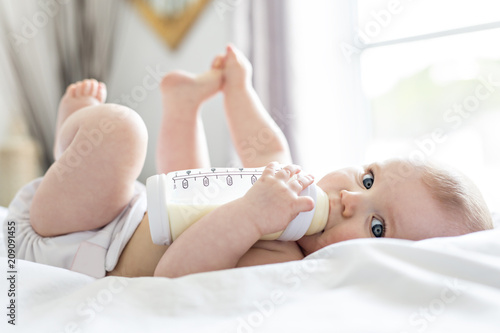 Pretty baby girl drinks water from bottle lying on bed. Child weared diaper in nursery room.