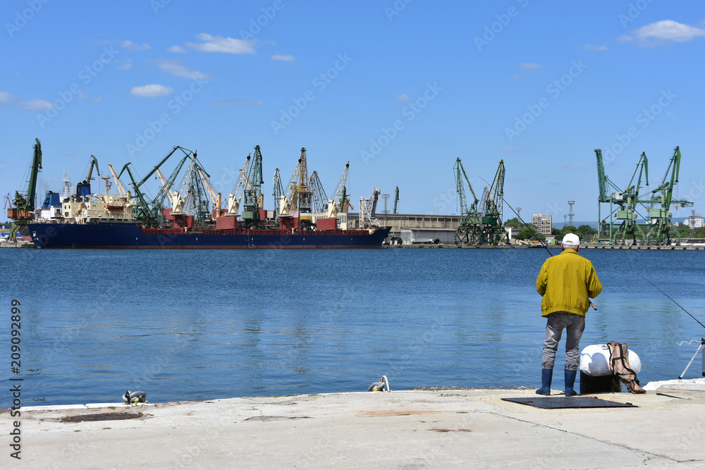 Varna Harbour, Bulgaria. Elderly fisherman fishing on the pier. Cranes and freighters seen at background. Varna Harbour, Bulgaria. Elderly fisherman fishing on the pier. Cranes and freighters seen at background.