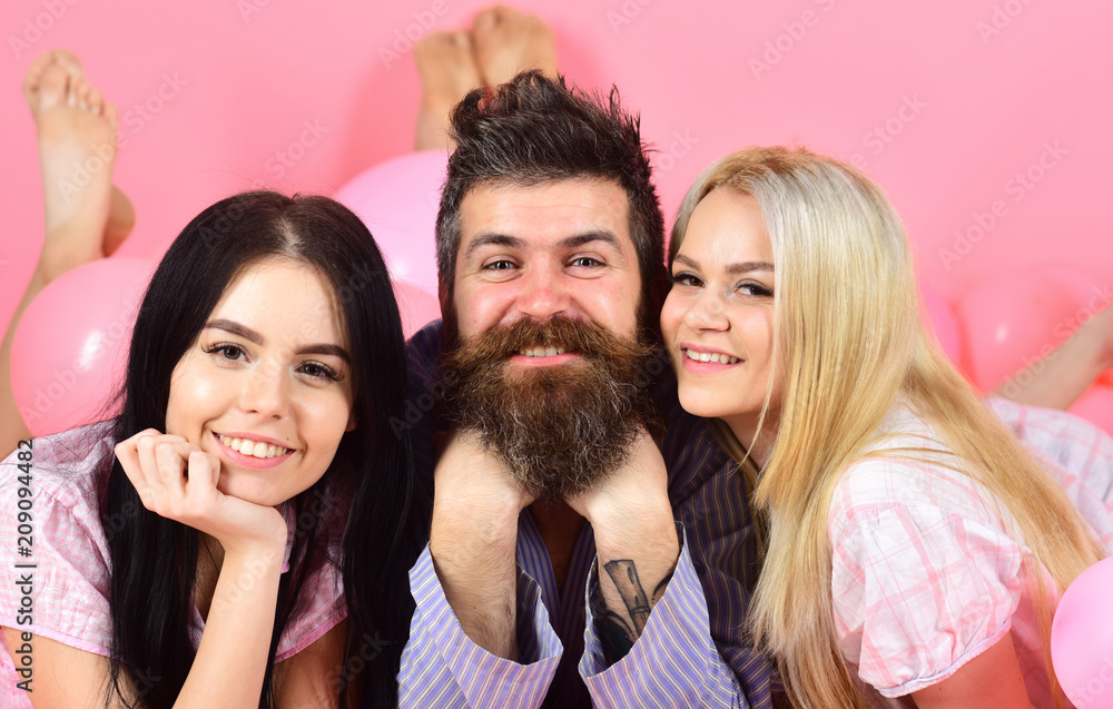 Man with beard and mustache attracts blonde and brunette girls. Girls fall in love with bearded macho, pink background. Threesome on smiling faces lay near balloons. Alpha male concept.