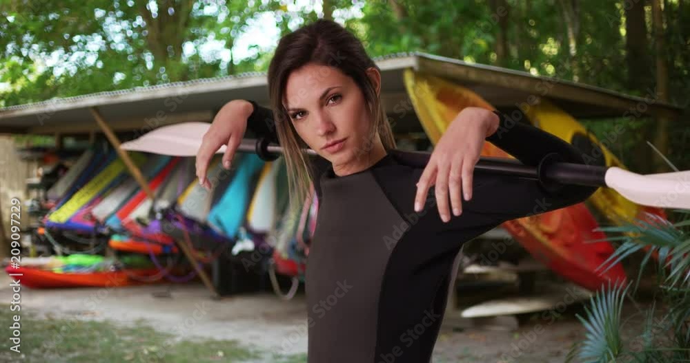 Young woman in wetsuit holding kayak paddle over shoulders near kayaks