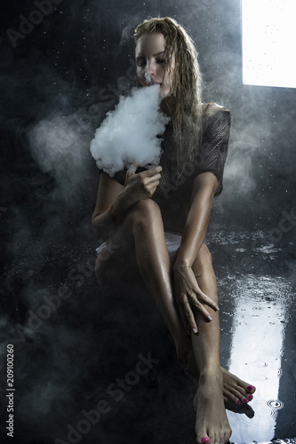 Beautiful topless athletic big breasted blonde girl wearing white sexy bodysuit and mesh transparent top vaping sitting on wet floor in scenic smoke under falling water drops on black.
