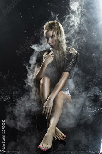 Beautiful topless athletic big breasted blonde girl wearing white sexy bodysuit and mesh transparent top vaping sitting on wet floor in scenic smoke under falling water drops on black.