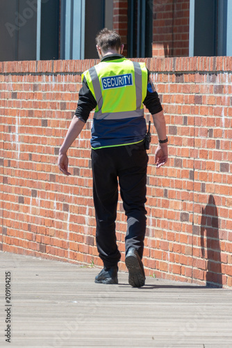 security guard in yellow hi vis with walkie taklie walking away from job on wooden decking by brick wall after being fired with head bowed