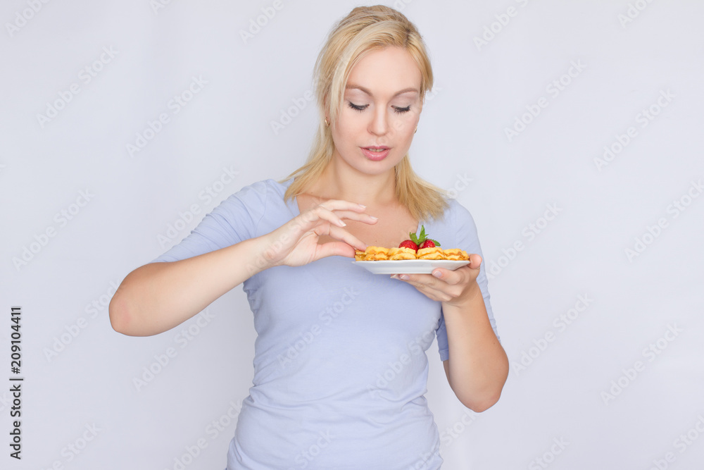 A happy blonde is holding a plate with Belgian waffles in her hands. Breakfast is decorated with strawberries and mint leaves. Concept: home food. A happy blonde is holding a plate with Belgian waffles in her hands. Breakfast is decorated with strawberries and mint leaves. Concept: home food.