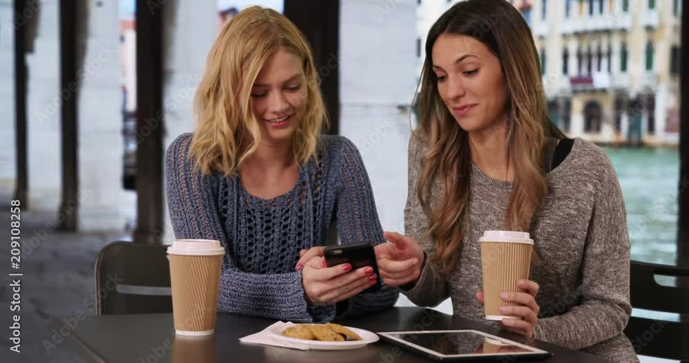 Two charming young friends using cell phone while sitting at table with tablet and drinks in Venice, Italy, Couple of attractive girls sitting and sharing pictures on smart phone by Venetian canal, 4k Two charming young friends using cell phone while sitting at table with tablet and drinks in Venice, Italy, Couple of attractive girls sitting and sharing pictures on smart phone by Venetian canal, 4k