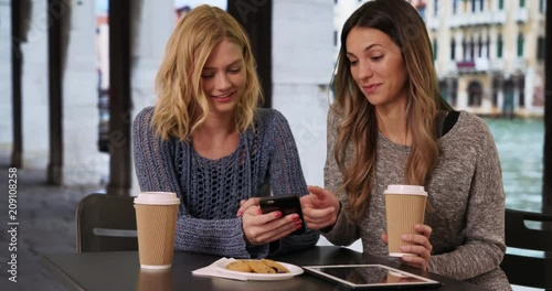 Two charming young friends using cell phone while sitting at table with tablet and drinks in Venice, Italy, Couple of attractive girls sitting and sharing pictures on smart phone by Venetian canal, 4k
