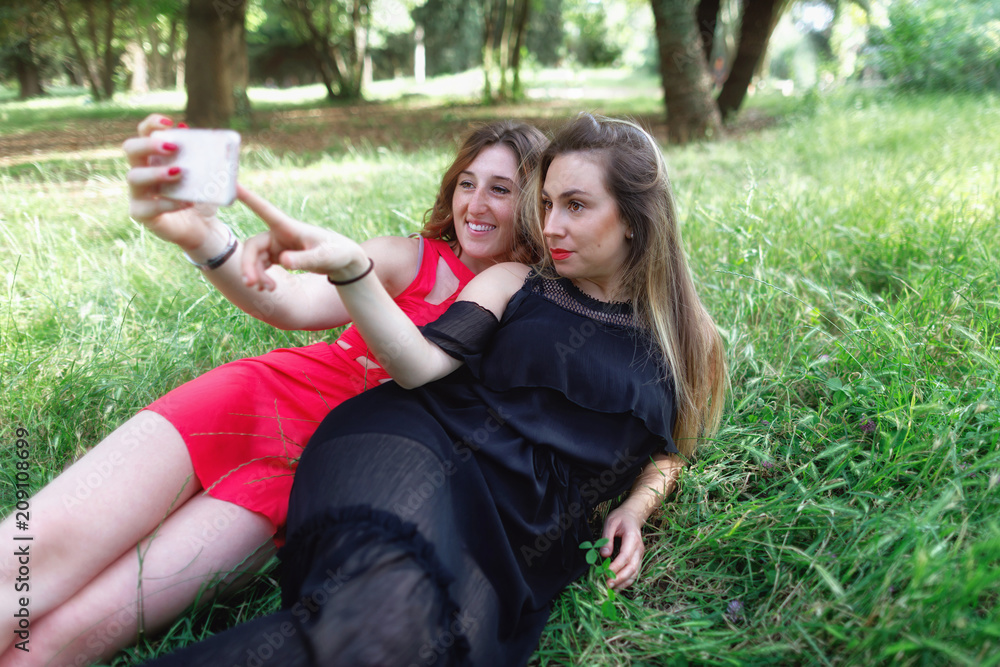 Girls smile and take a selfie together. Two girls stretched out in the grass take a selfie together and smile. In the background the green in the outdoor park.