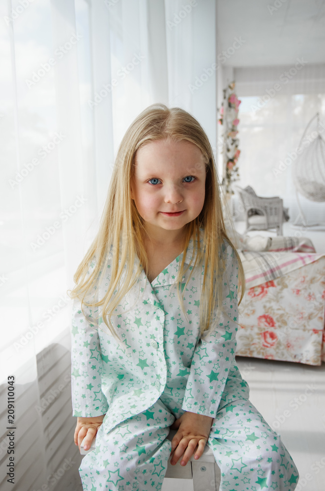 A little blonde girl sits on a chair by the window. Girl in pajamas.