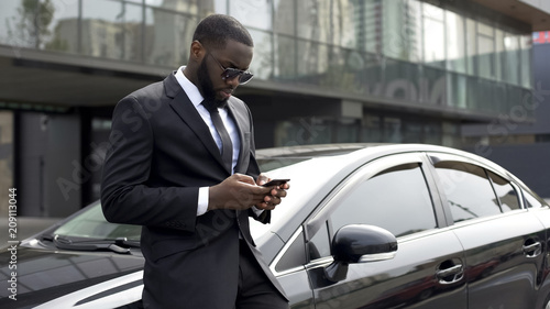 Confident man typing e-mail on smartphone, standing near his luxury car © motortion