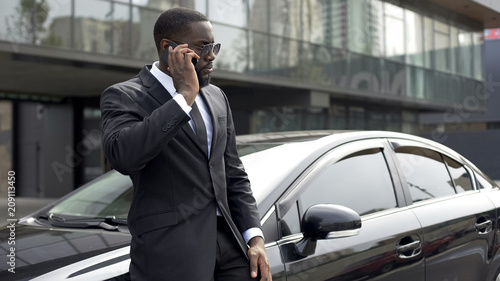 Manager in expensive suit speaking on phone with customer, standing near his car