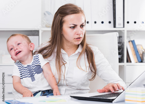 Girl is working with a naughty child in her arms behind a laptop