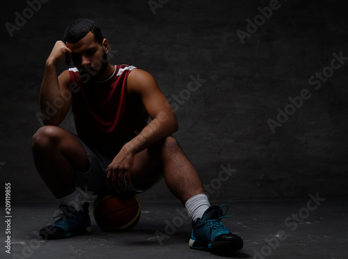 Pensive young African-American basketball player in sportswear sitting on a ball over dark background.