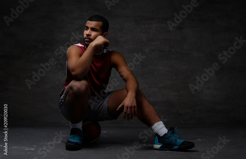 Pensive young African-American basketball player in sportswear sitting on a ball over dark background.