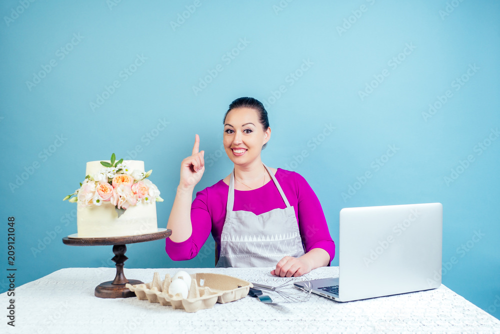 confectioner housewife (pastry-cook) woman points finger to the up next to creamy white two-tiered wedding cake with fresh flowers and a laptop on a table in studio on a blue background copyspace confectioner housewife (pastry-cook) woman points finger to the up next to creamy white two-tiered wedding cake with fresh flowers and a laptop on a table in studio on a blue background copyspace