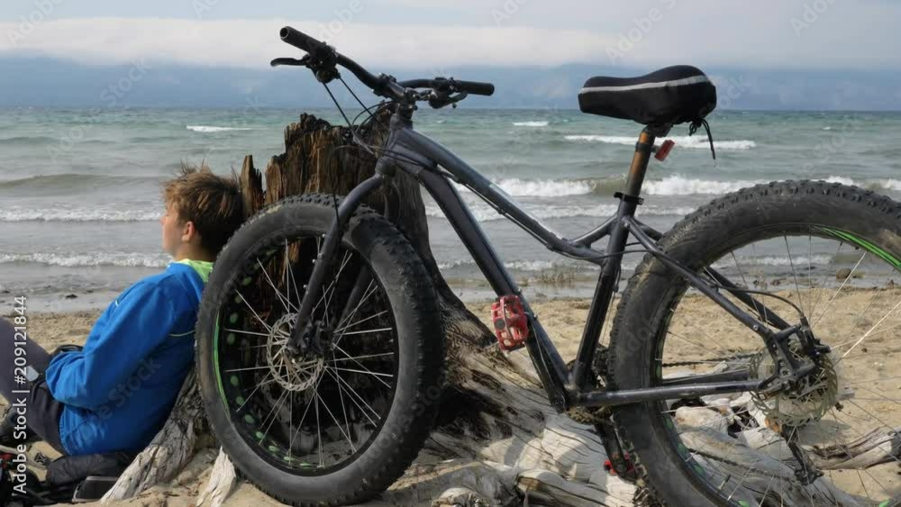 Fat bike also called fatbike or fat-tire bike in summer driving on the beach. The guy and his bike rest on the sandy beach. He sits and rests after a long trip. Enjoys the beautiful sea view and the Fat bike also called fatbike or fat-tire bike in summer driving on the beach. The guy and his bike rest on the sandy beach. He sits and rests after a long trip. Enjoys the beautiful sea view and the