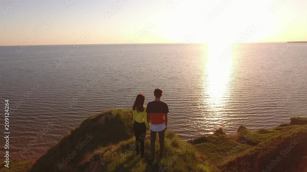 A bird`s eye zoom in of a young couple in love standing on a peak and watching the golden sun path at the Black Sea at sunset in summer A bird`s eye zoom in of a young couple in love standing on a peak and watching the golden sun path at the Black Sea at sunset in summer