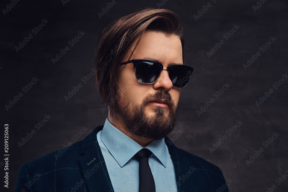 Close-up portrait of a handsome fashionable middle-aged man with beard and hairstyle dressed in an elegant formal suit and sunglasses. Isolated on a textured dark background in studio. Close-up portrait of a handsome fashionable middle-aged man with beard and hairstyle dressed in an elegant formal suit and sunglasses. Isolated on a textured dark background in studio.